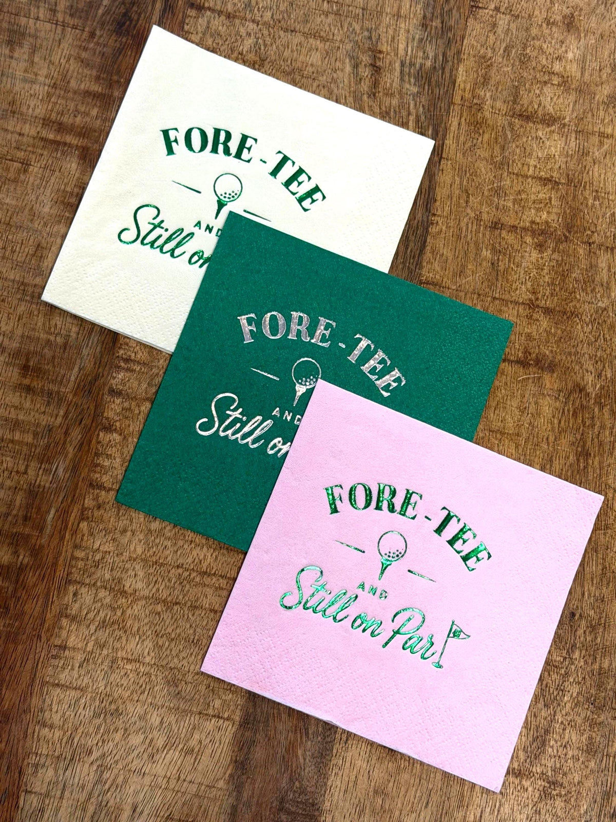 Three paper cocktail napkins in white, green, and pink are fanned out on a wooden surface, each printed with foil text reading FORE-TEE AND Still on Par alongside a golf ball on a tee and a flag.