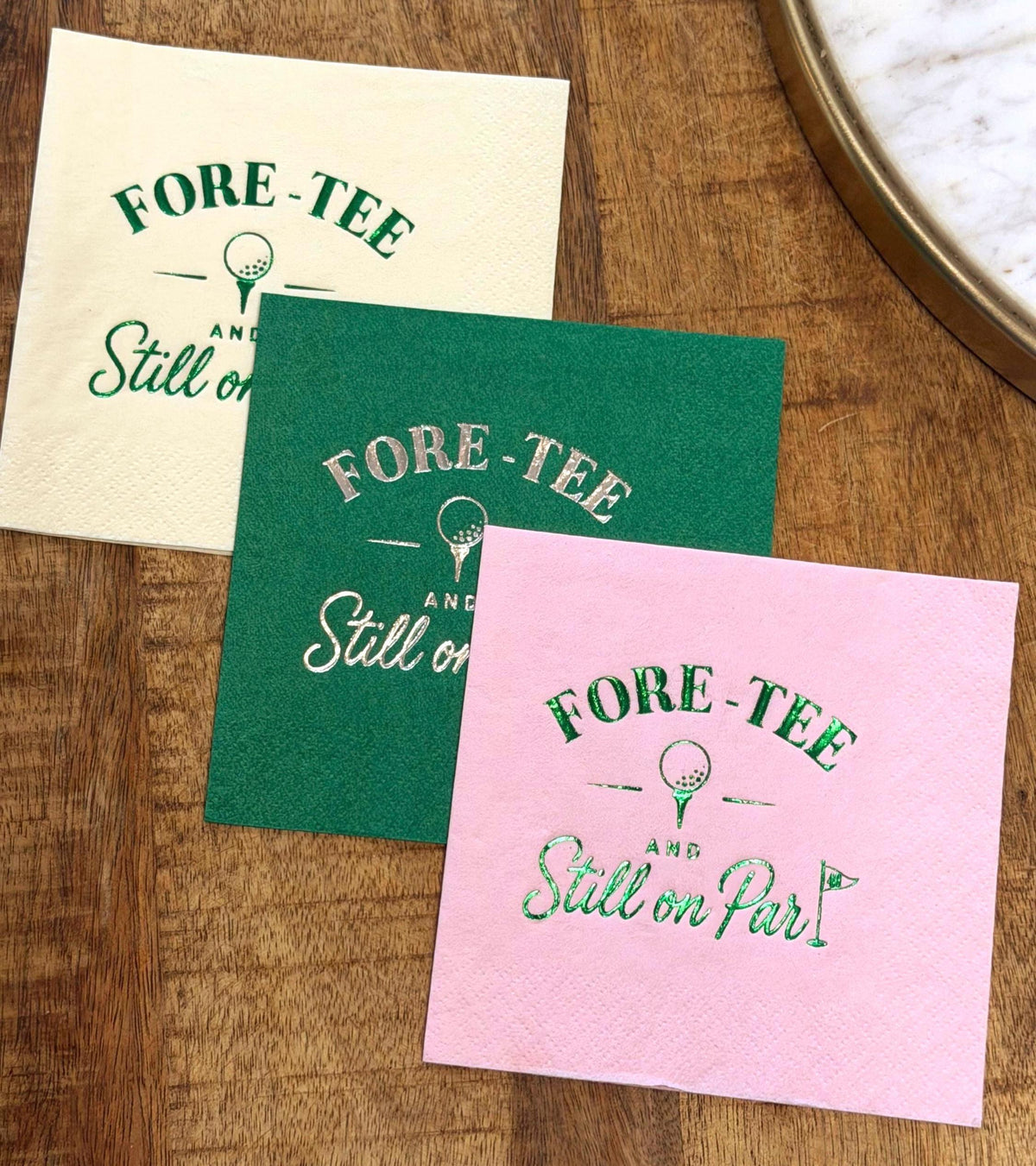 A stack of three layered cocktail napkins in cream, green, and pink sits on a wood background, displaying matching foil golf themed graphics and text.