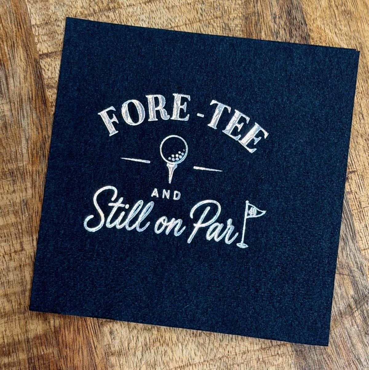 A solid black paper cocktail napkin on a wood surface features silver foil lettering that reads FORE-TEE AND Still on Par with a golf ball and flag motif.