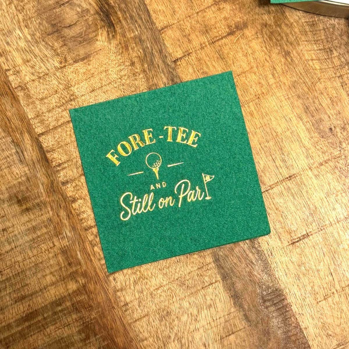 A green paper cocktail napkin lies on a wooden surface, stamped with gold foil text that says FORE-TEE AND Still on Par with golf illustrations.