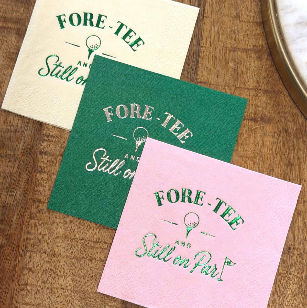 White, green, and pink cocktail napkins with golf-themed foil stamping sit on a wooden table surface.