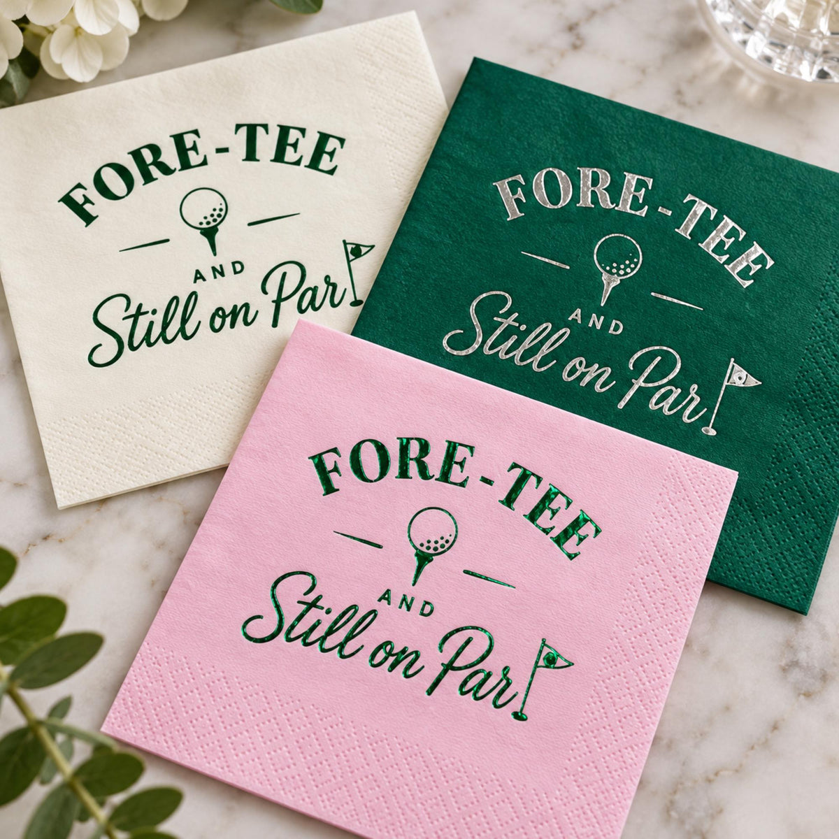 Three folded cocktail napkins in white, green, and pink display foil-stamped golf graphics and text on a marble background.