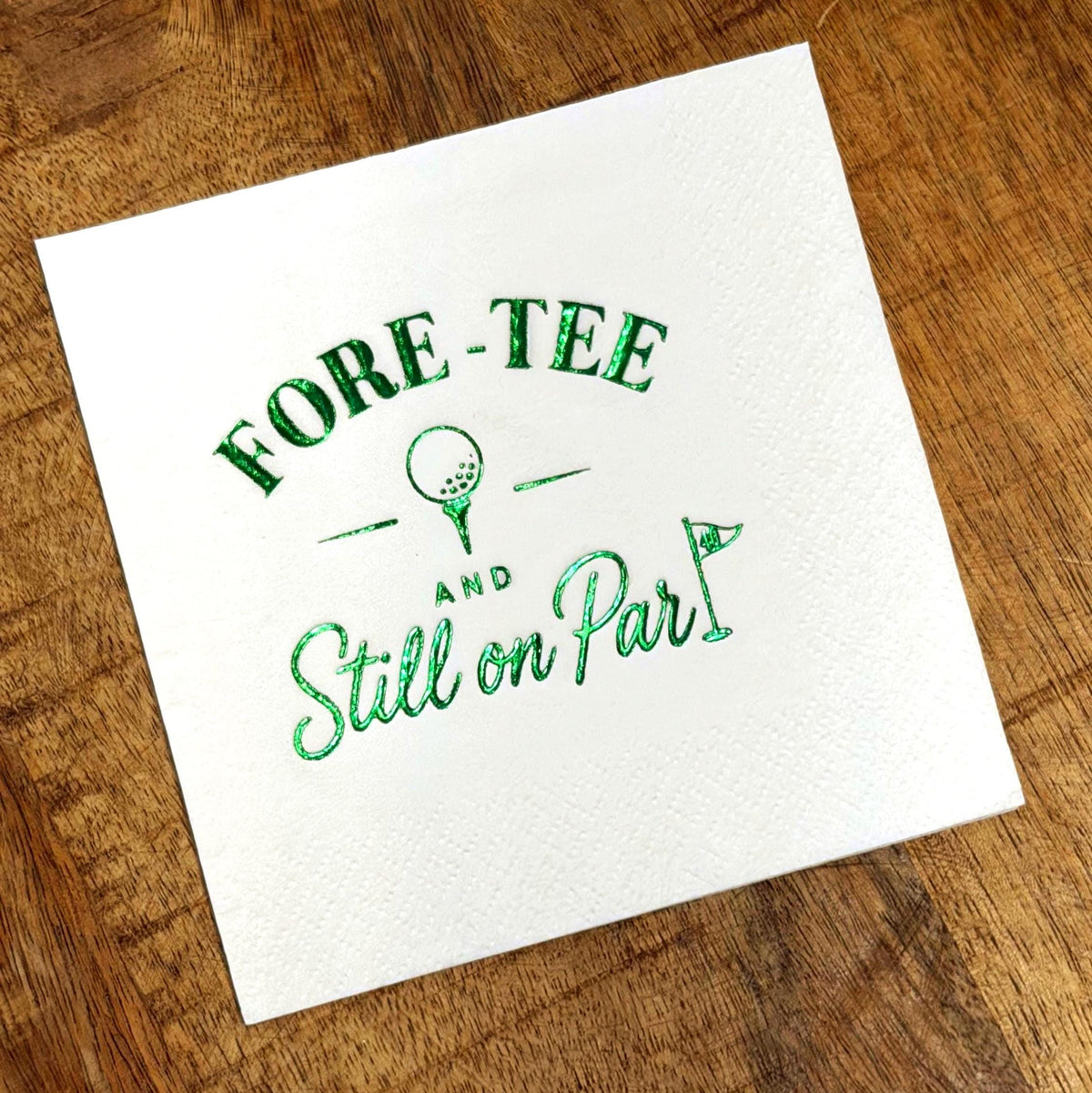 A single white cocktail napkin features green foil stamping with a golf ball on a tee and text on a wooden surface.