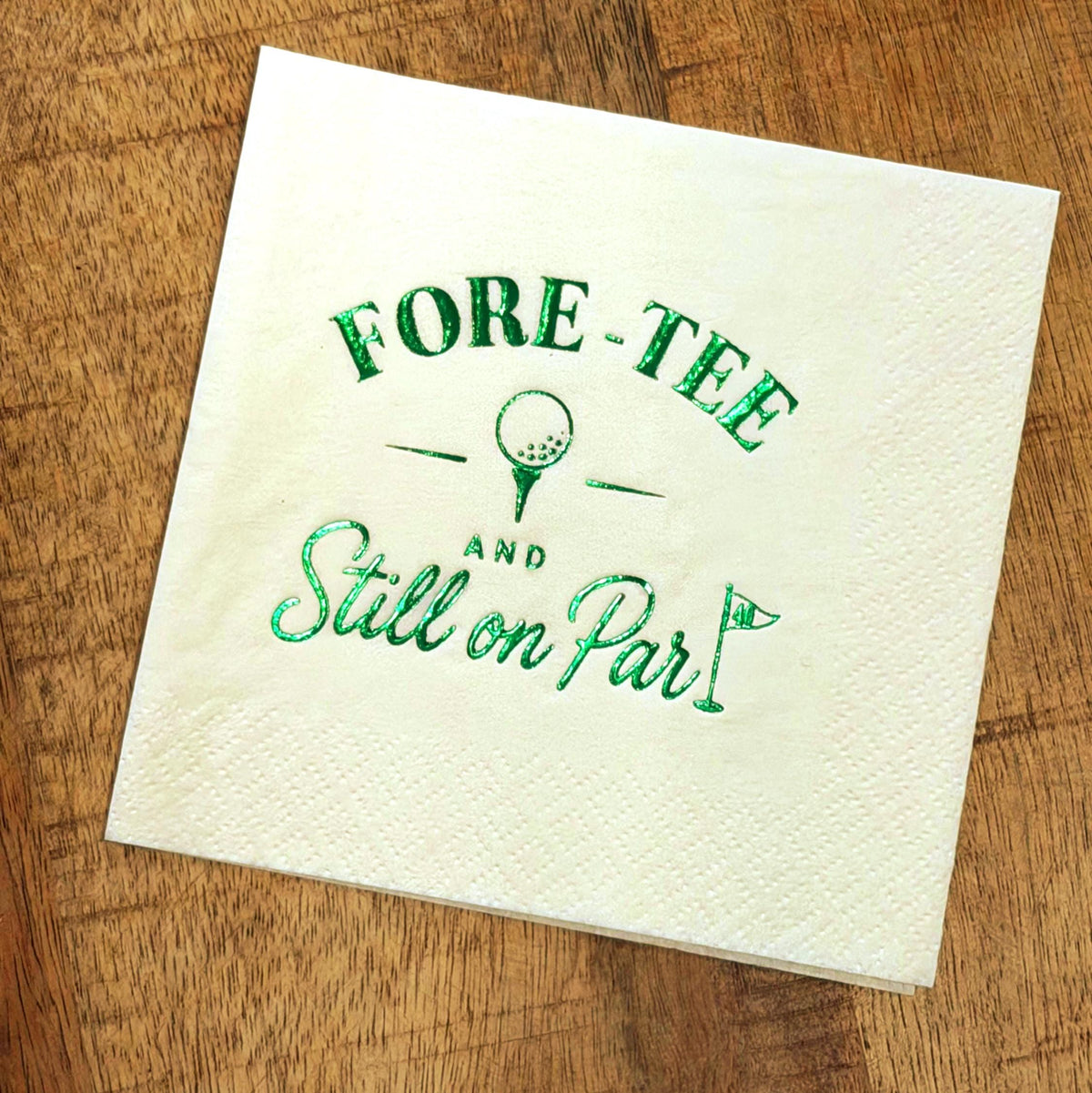 A white cocktail napkin printed with green foil golf text and graphics rests on a wooden table.