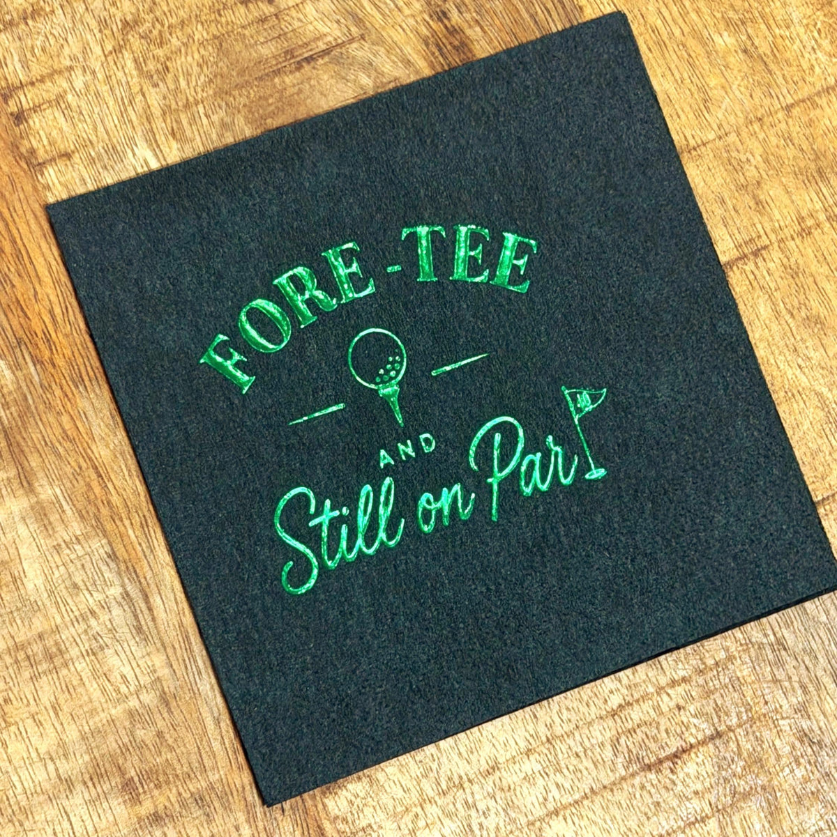 A dark green cocktail napkin features silver foil stamping of golf text and a golf ball graphic on a wooden surface.