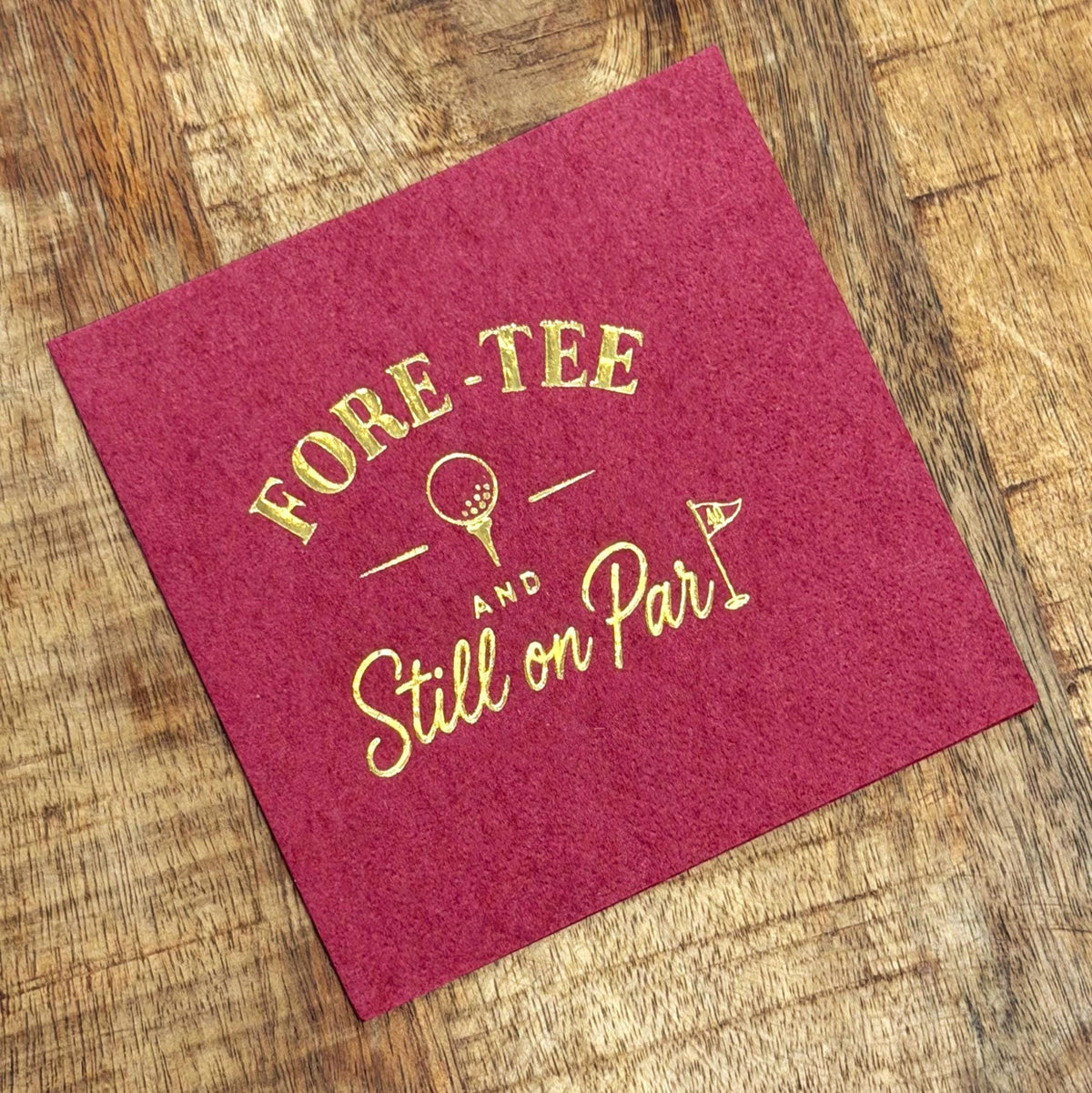 A pink cocktail napkin with gold foil stamping of a golf graphic and text lies on a wooden surface.