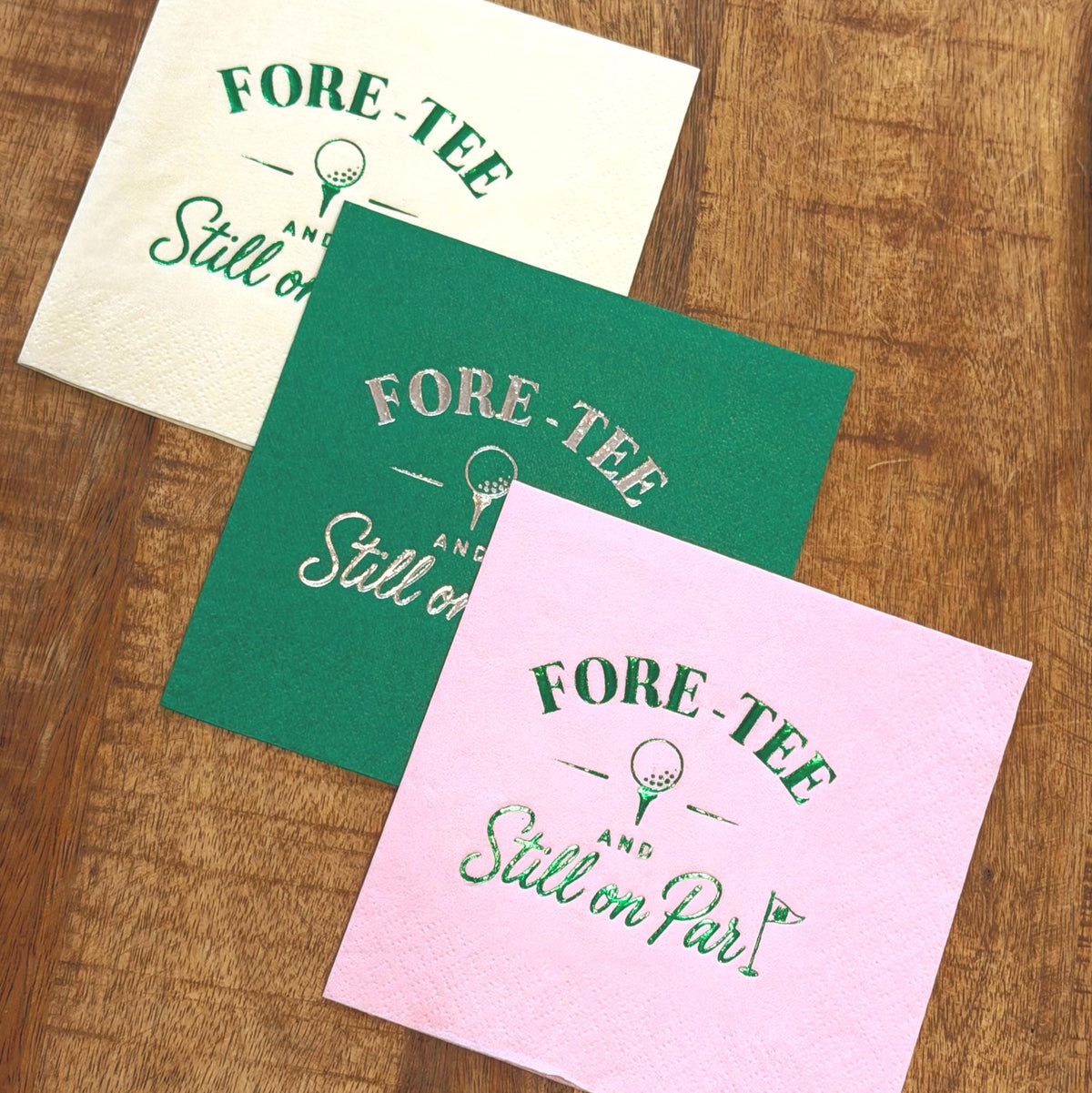 Three cocktail napkins in white, green, and pink featuring foil-stamped golf designs are arranged diagonally on a wooden tabletop.