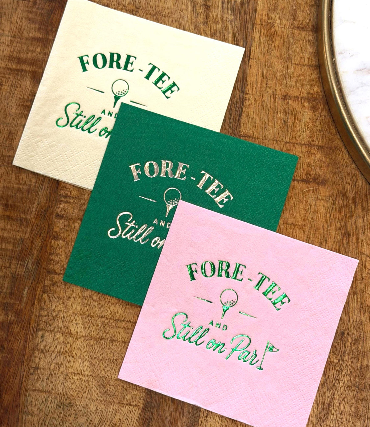 White, green, and pink cocktail napkins with foil-stamped golf themes are stacked diagonally on a wood surface next to a mirror edge.