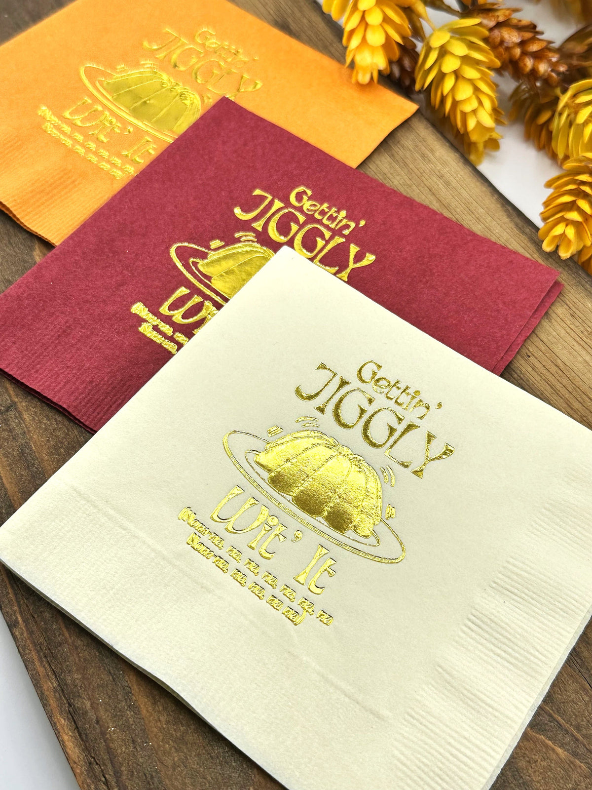 Three paper napkins with gold foil lettering. The napkins are in orange, red, and white. The text on the napkins reads "Gettin' Jiggly" and "Wit It".