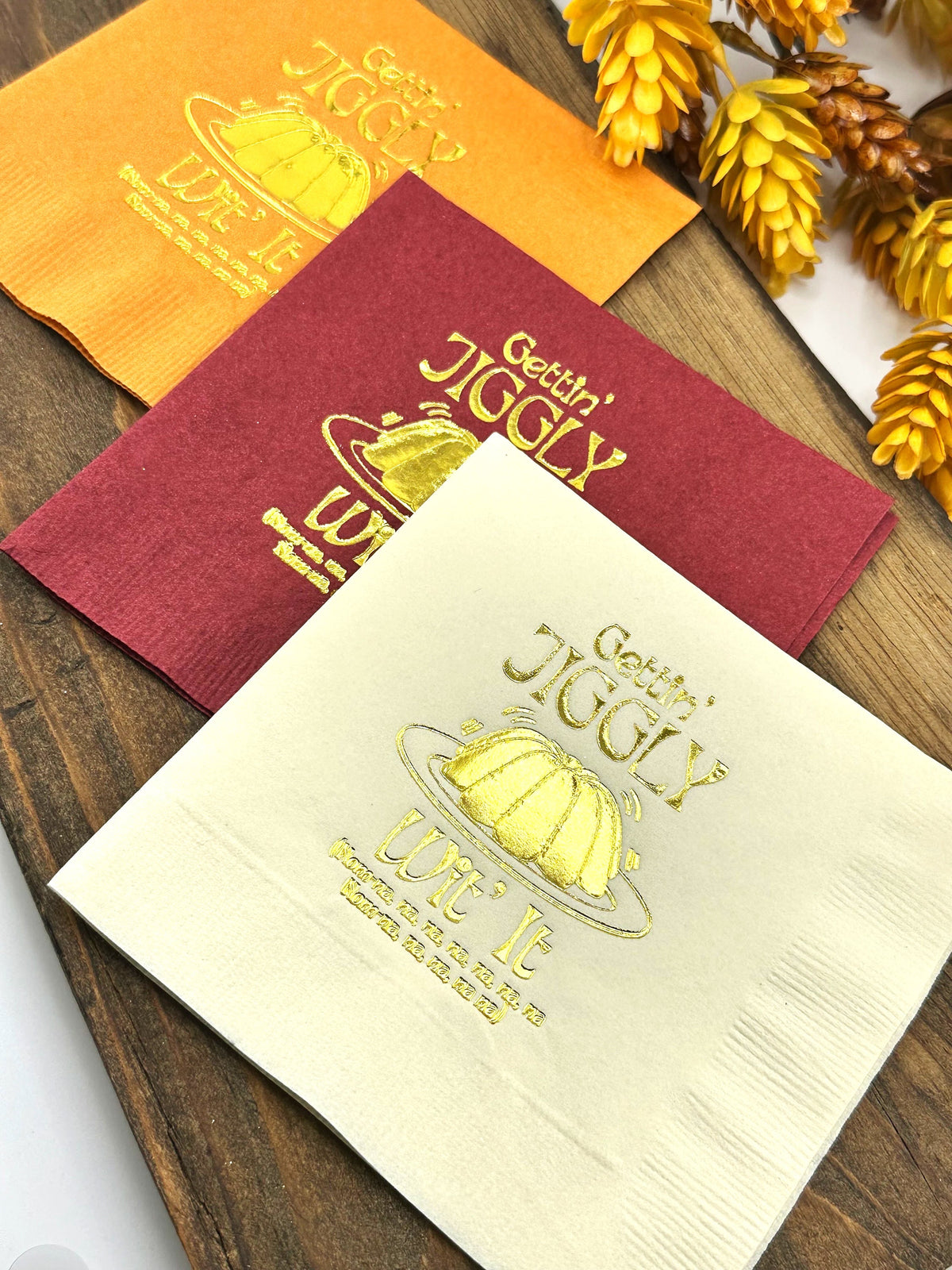 Three napkins with a gold design of a plate of jiggly dessert with the text "Gettin' Jiggly" and "Wob It" on a wooden surface.