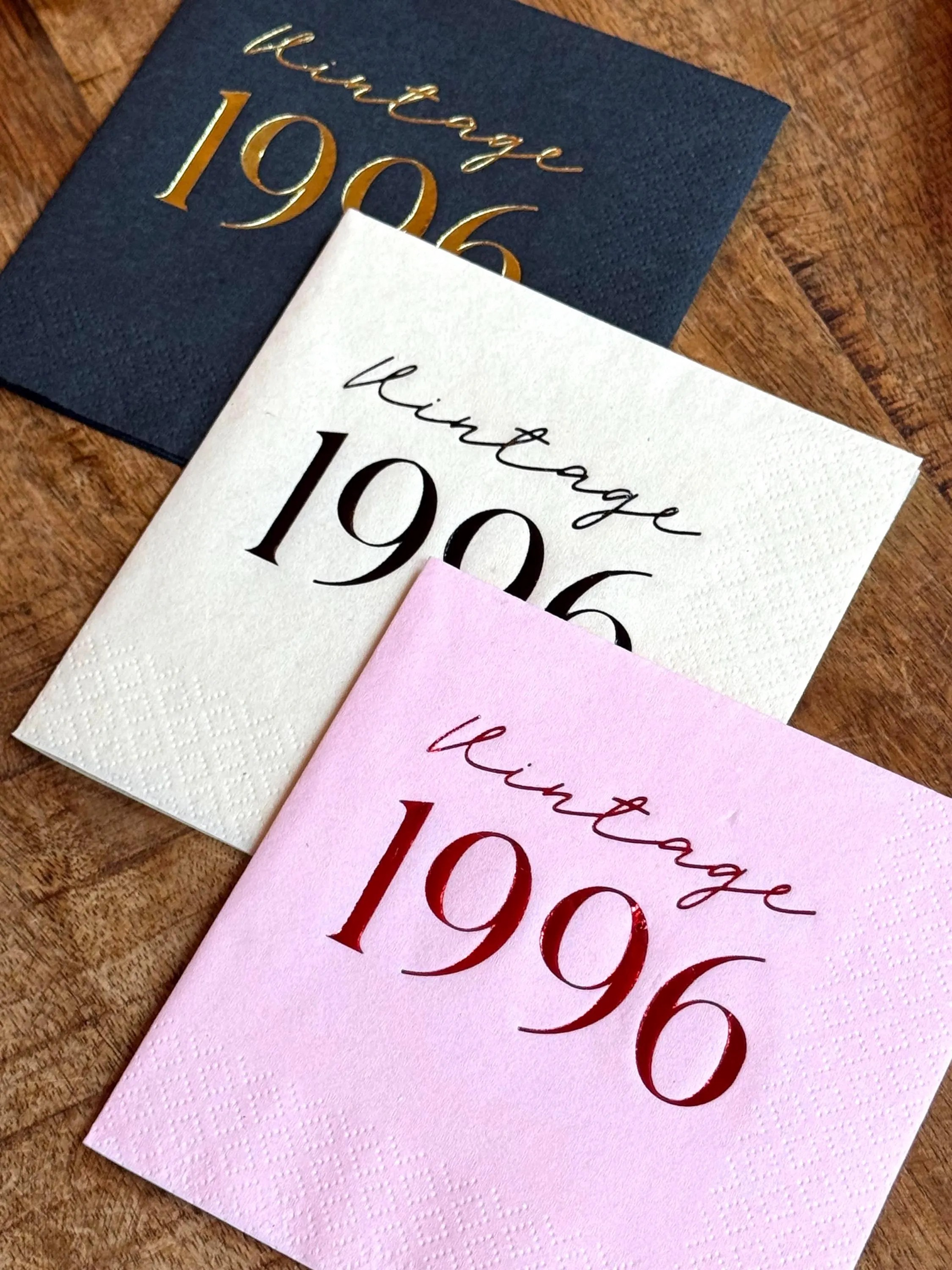 Three cocktail napkins with 'Vintage 1996' text in different colors on a wooden surface.