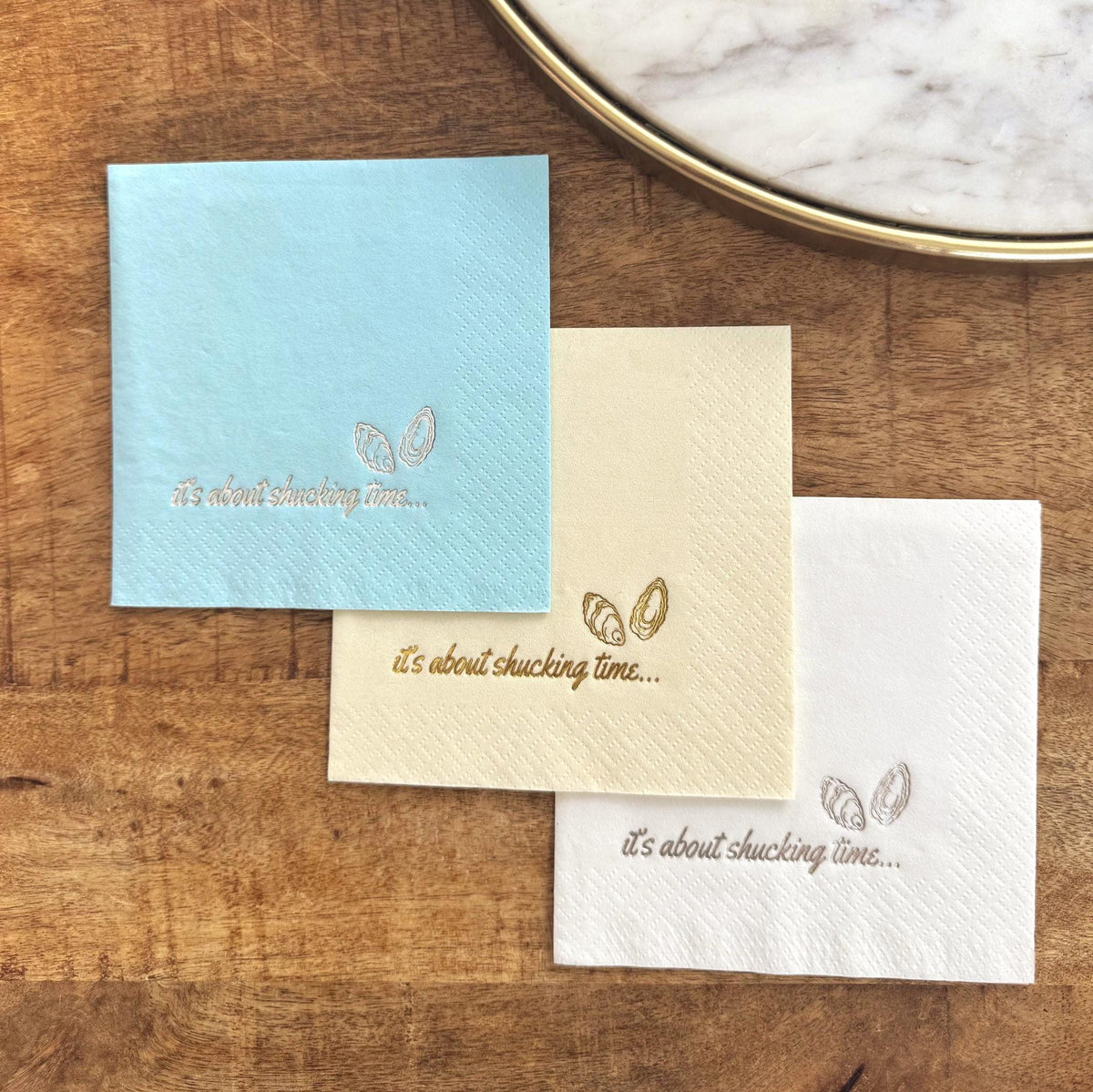 Three square paper napkins in light blue, cream, and white, each with the phrase "it's about shucking time..." and an oyster illustration. The napkins are arranged on a wooden surface, suggesting a coastal or seafood theme.