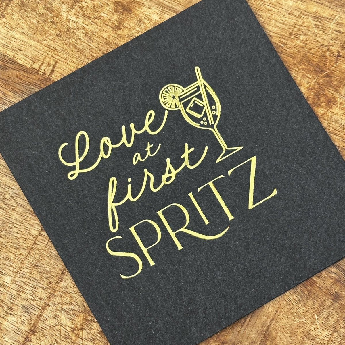 A square black cocktail napkin with gold foil stamping reading Love at first SPRITZ and a cocktail glass graphic resting on a wooden surface.