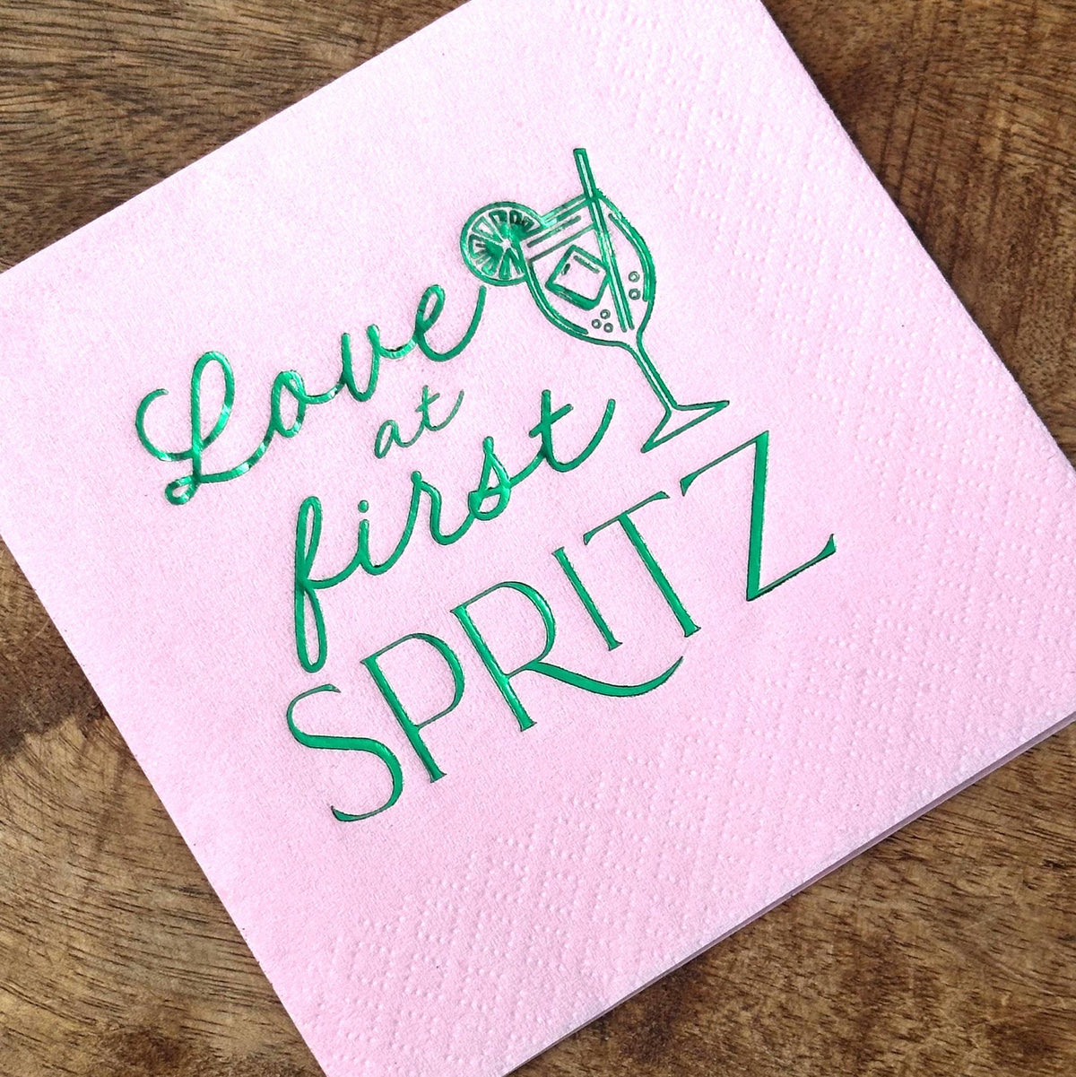 A square light pink cocktail napkin featuring shiny green foil text reading Love at first SPRITZ with a cocktail glass graphic placed on a wooden table.