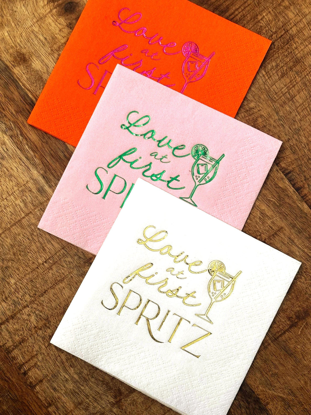 Three folded cocktail napkins in orange, pink, and white on a wooden surface, each printed with the Love at First Spritz design.