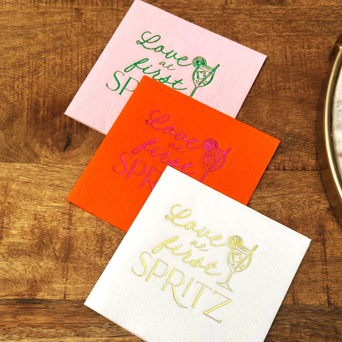 Three cocktail napkins in pink, orange, and white stacked on a wooden background with matching drink graphics.