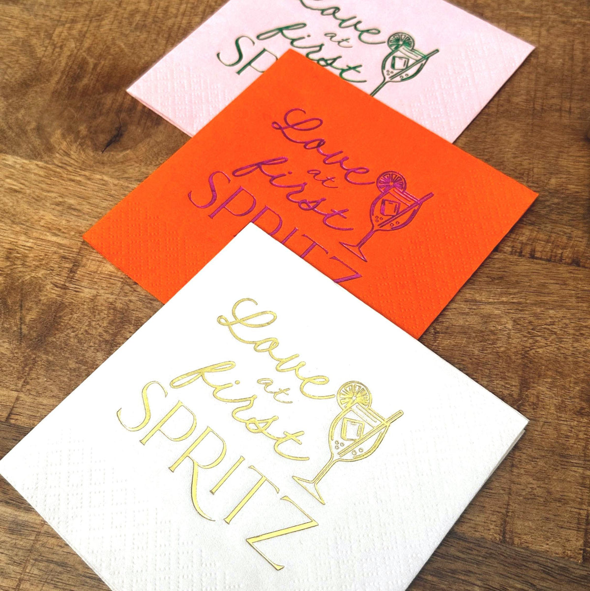 Three layered cocktail napkins in pink, orange, and white on a wood table, featuring foil stamped text and a cocktail glass.