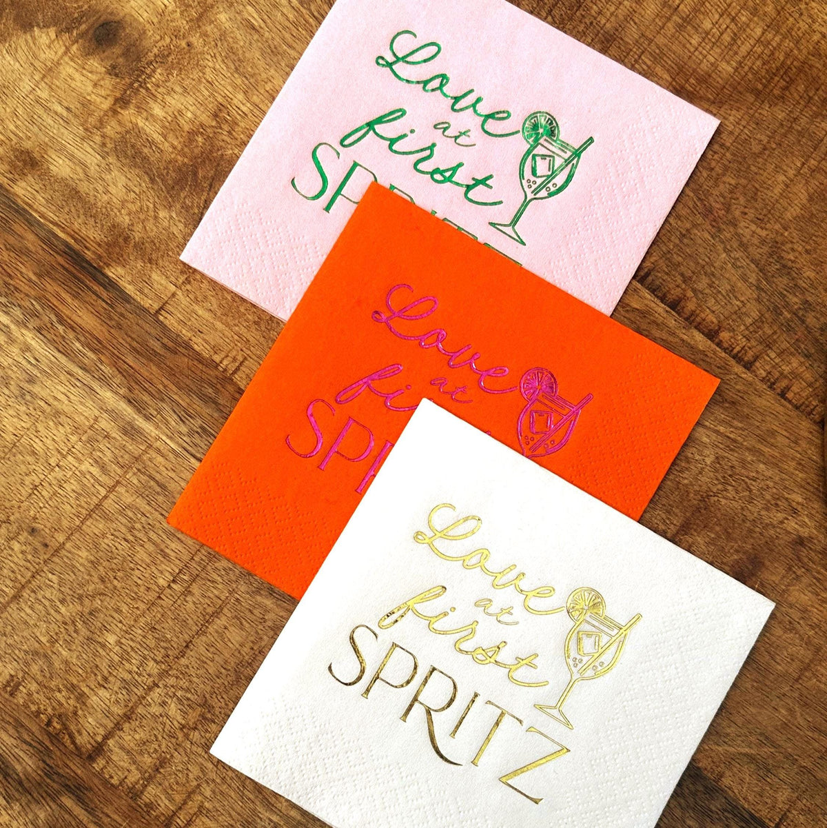 Three folded cocktail napkins in pink, orange, and white arranged diagonally on a wooden tabletop.