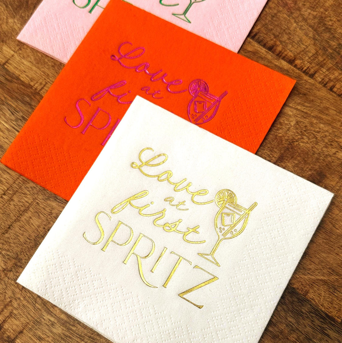 Three paper cocktail napkins in orange, pink, and white stacked on a wooden table, featuring colorful foil stamped cocktail graphics.