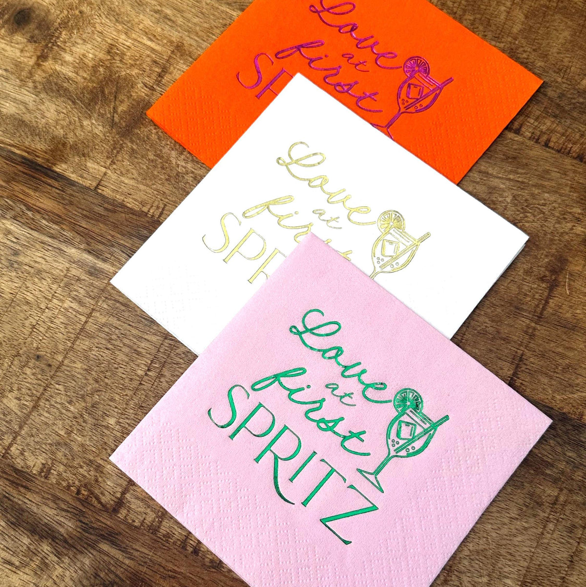 Three cocktail napkins in orange, white, and pink arranged on a wooden surface with foil stamped designs.