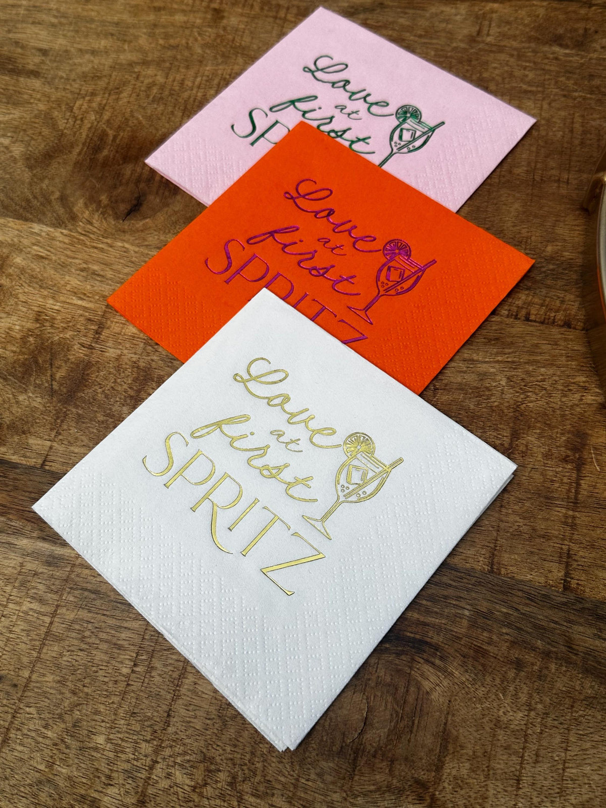 Three folded cocktail napkins in pink, orange, and white stacked diagonally on a wood surface.