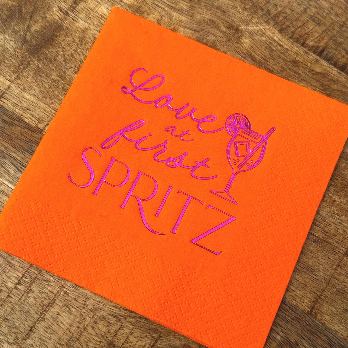 A single bright orange cocktail napkin on a wooden background, featuring purple foil stamping of a cocktail and the words Love at First Spritz.