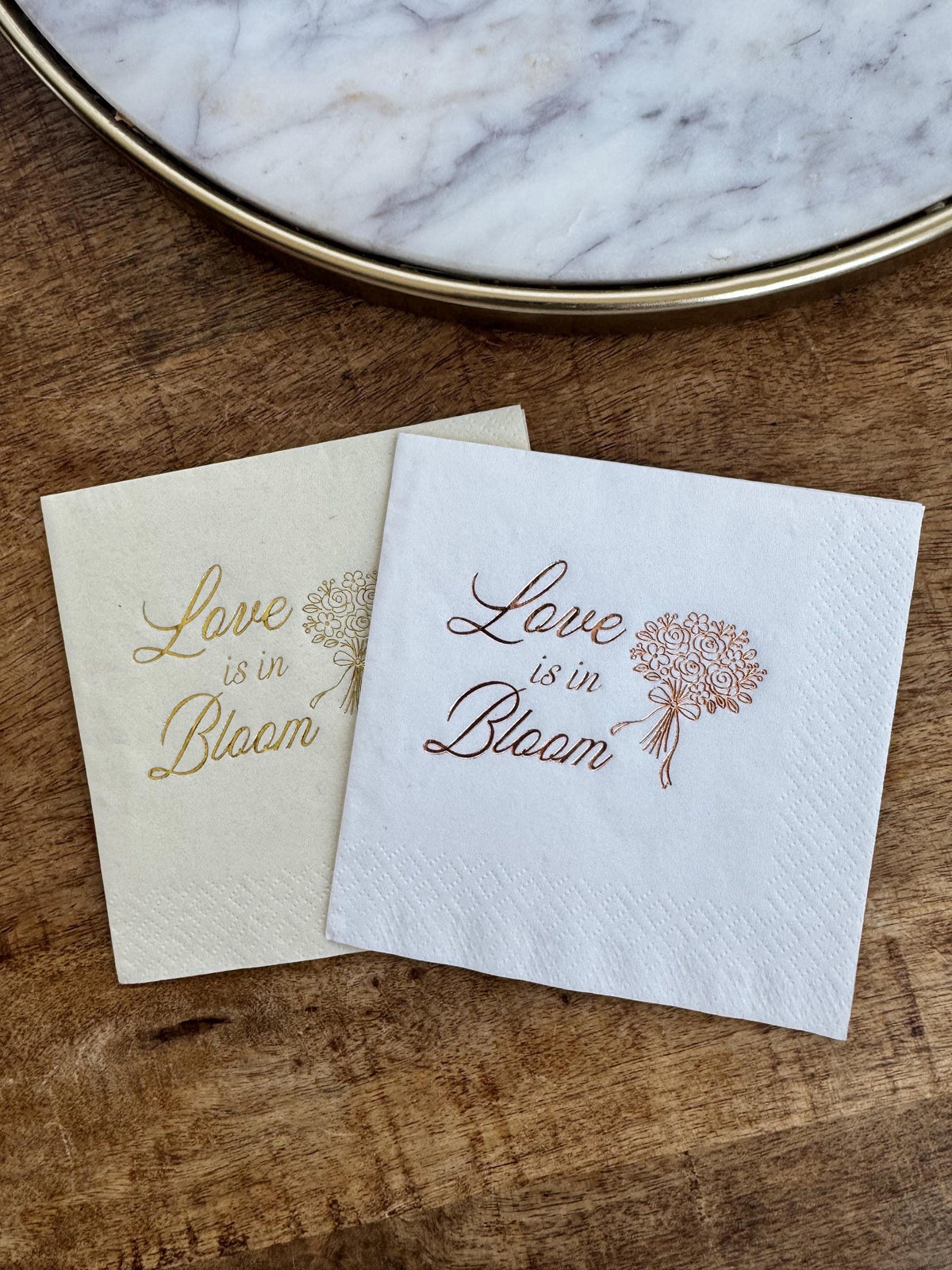 Two square napkins, one cream with gold print and one white with copper print, arranged on a dark wood table.