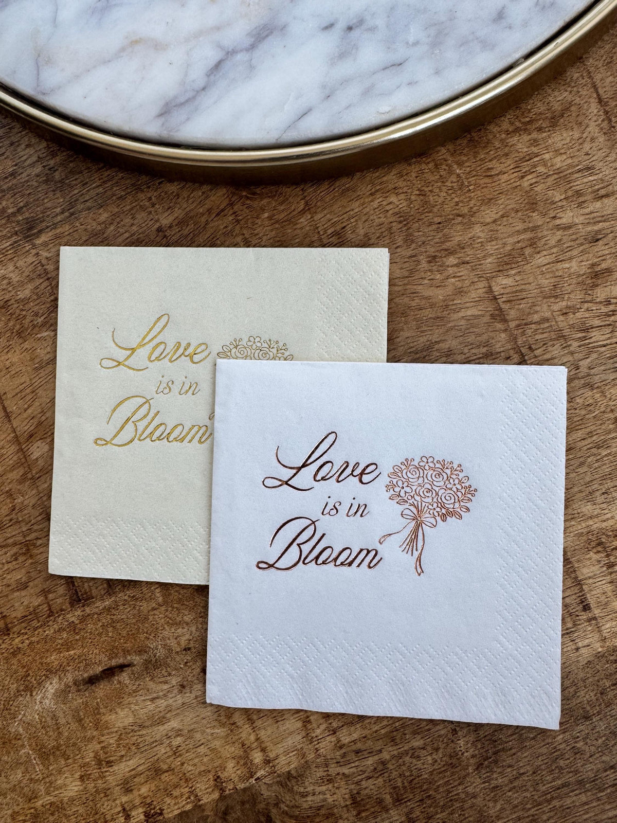 A cream napkin with gold lettering and a white napkin with copper lettering placed diagonally on a wooden tabletop.