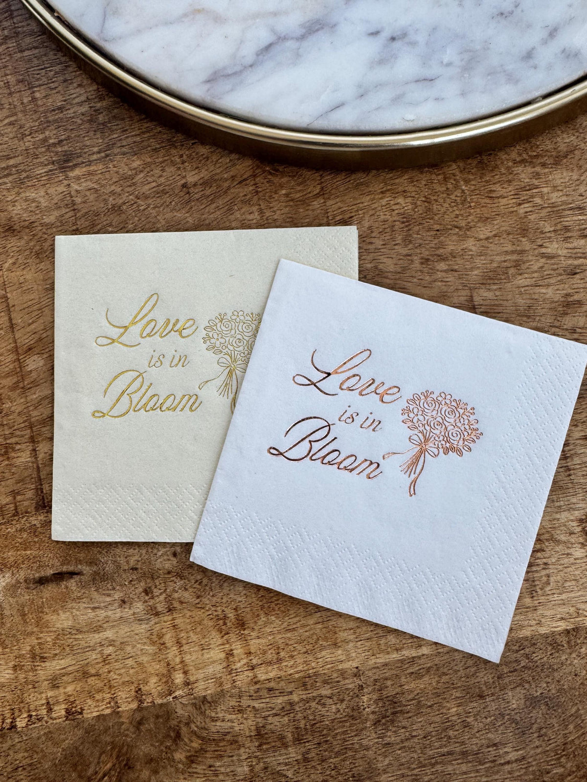 A cream napkin with light gold foil and a white napkin with copper foil resting on a wooden surface beneath a marble tray.