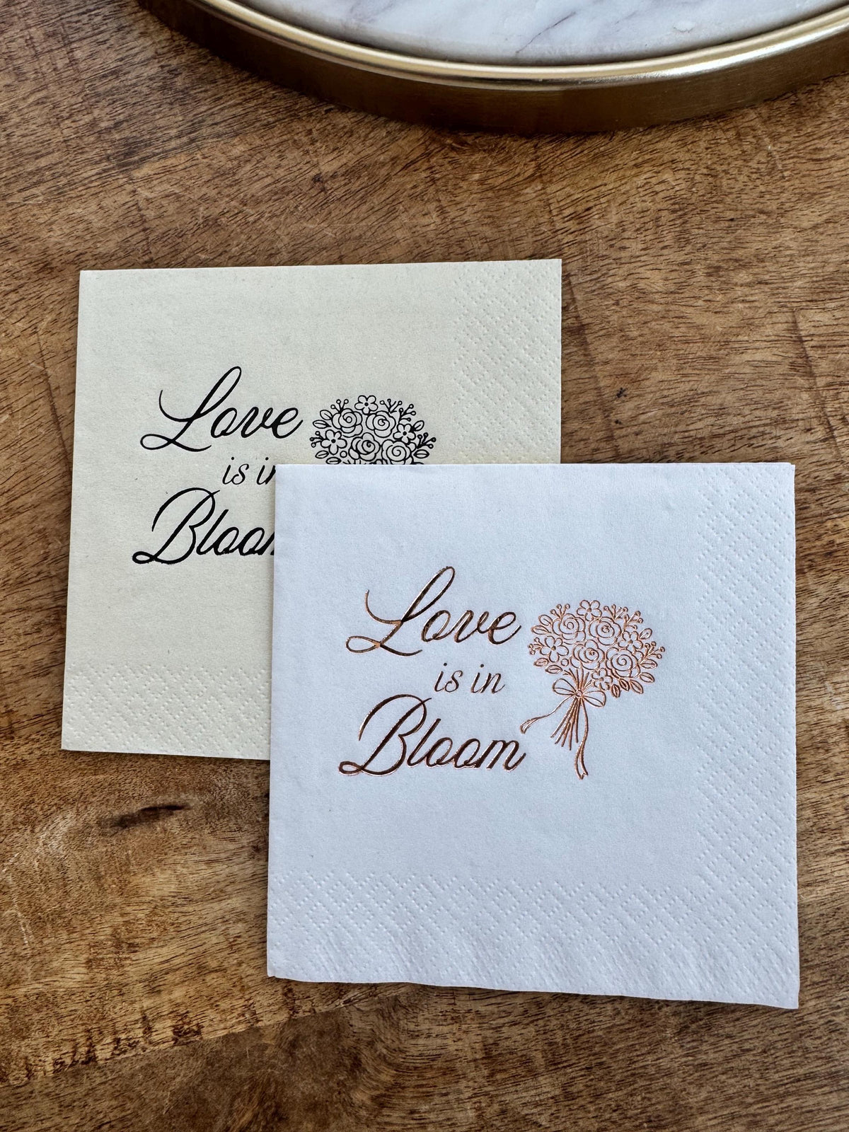 A cream napkin with black print overlapping a white napkin with copper print on a wooden board.