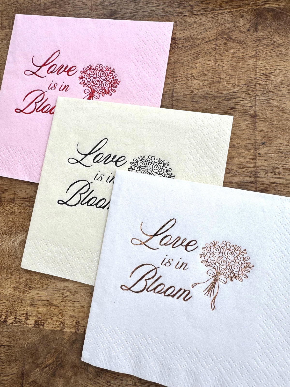 Three cocktail napkins stacked on a wooden background in pink with red foil, cream with black foil, and white with copper foil.