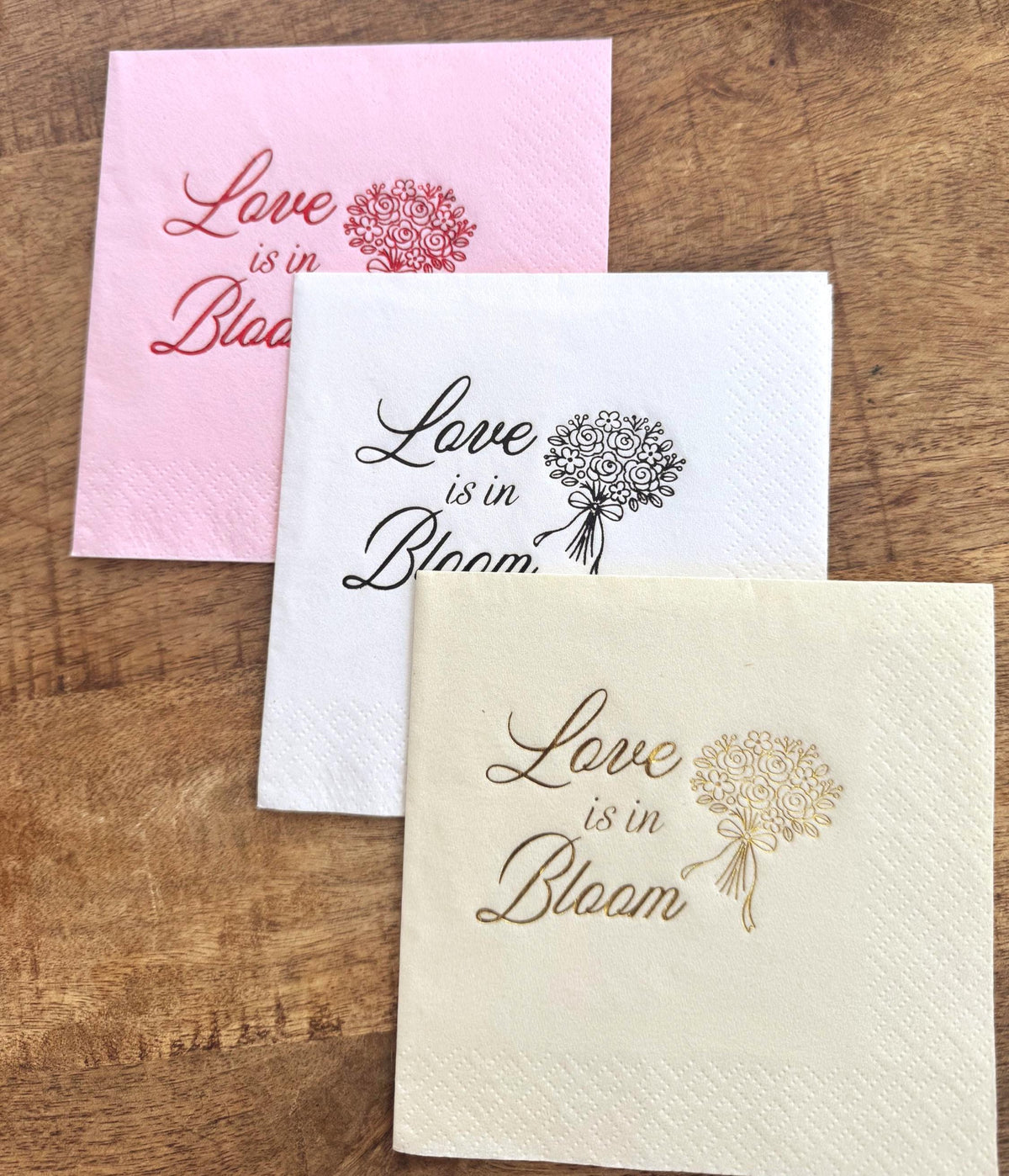 Three folded paper cocktail napkins stacked diagonally on a wooden surface, featuring light pink, white, and cream colors with varying foil colors of the text Love is in Bloom and a floral bouquet.