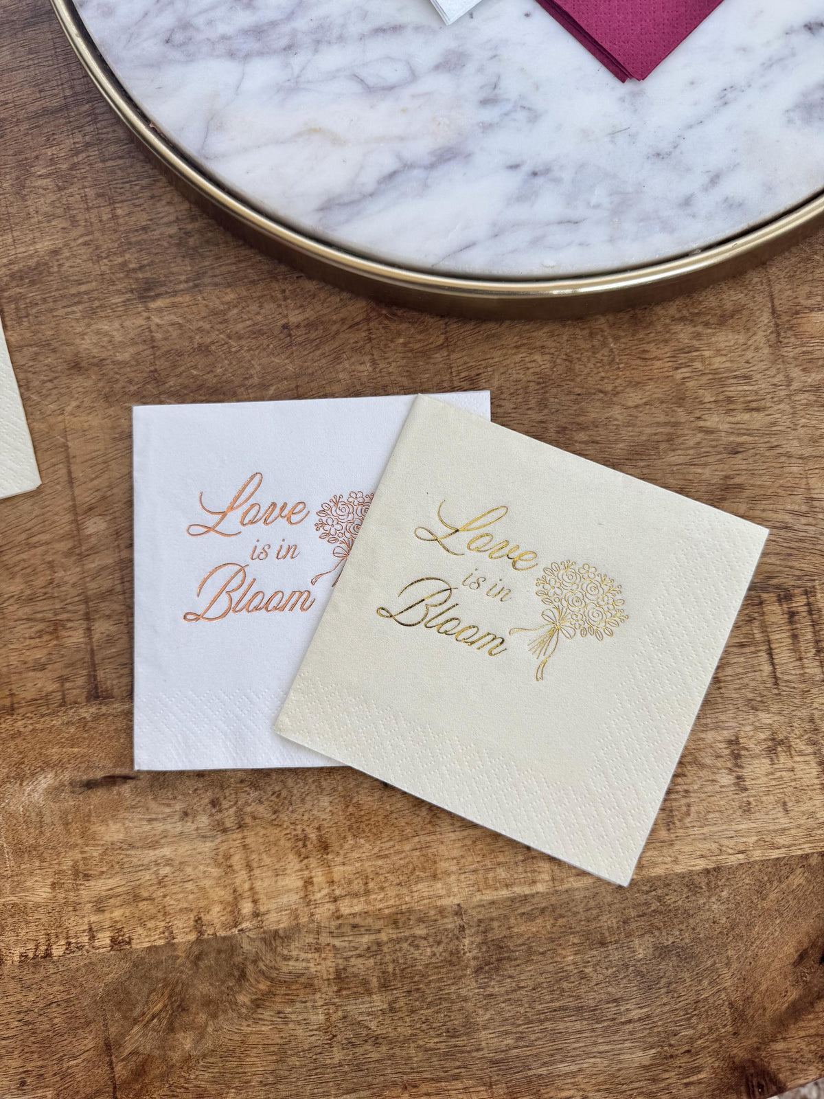 Two square paper cocktail napkins in white and cream lie on a wooden table beside a marble tray, printed with rose gold foil text reading Love is in Bloom alongside a tied flower bouquet.