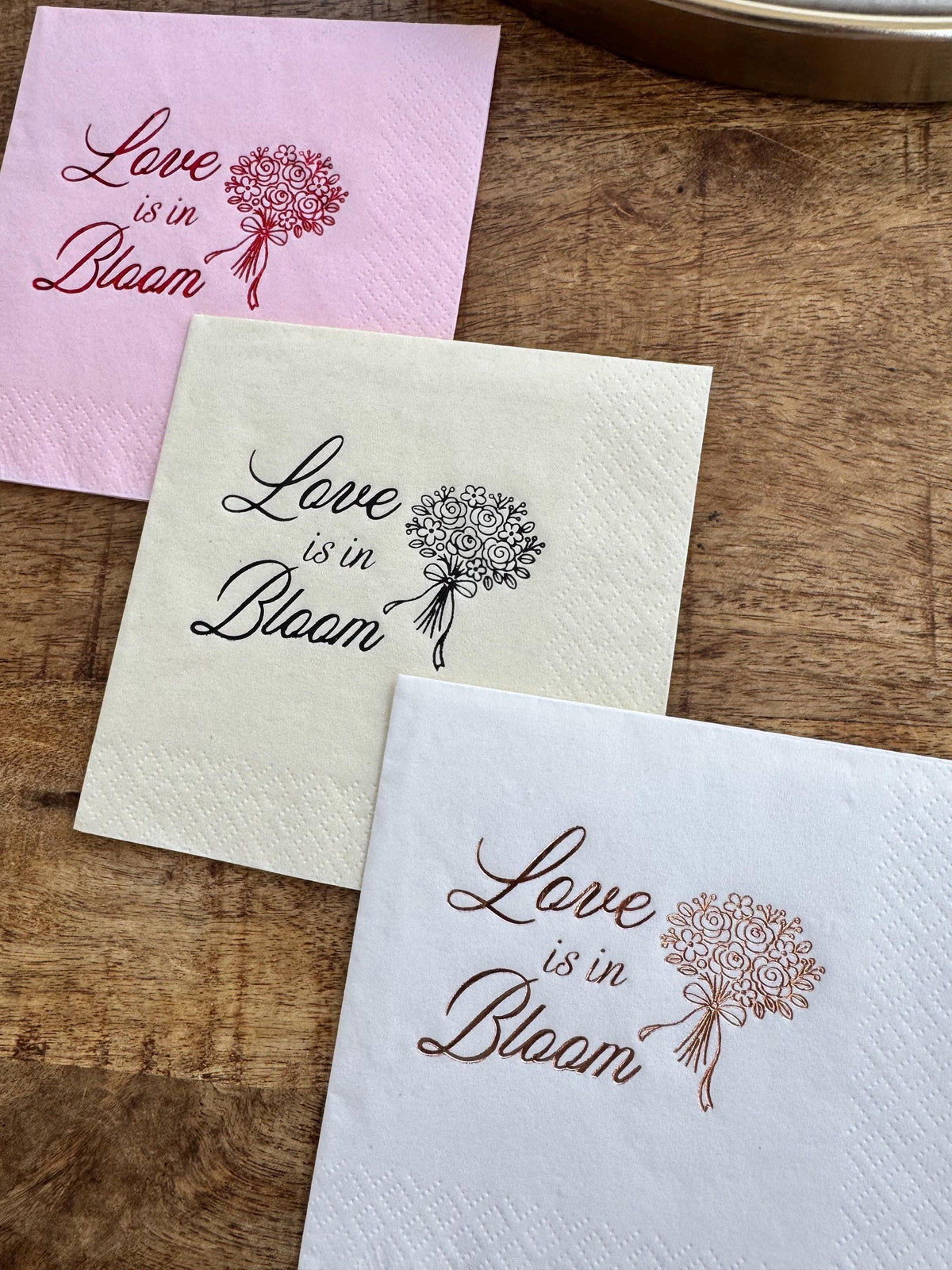 Three paper cocktail napkins in pink, cream, and white are arranged overlapping on a wooden surface, each printed with the text Love is in Bloom and a bouquet illustration in different foil colors.