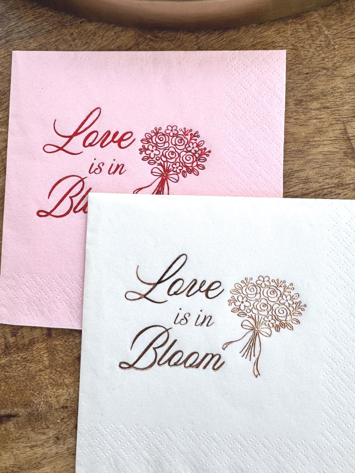 Two paper cocktail napkins on a wooden surface, a pink one with red foil print and a white one with copper foil print that reads Love is in Bloom with a bouquet illustration.