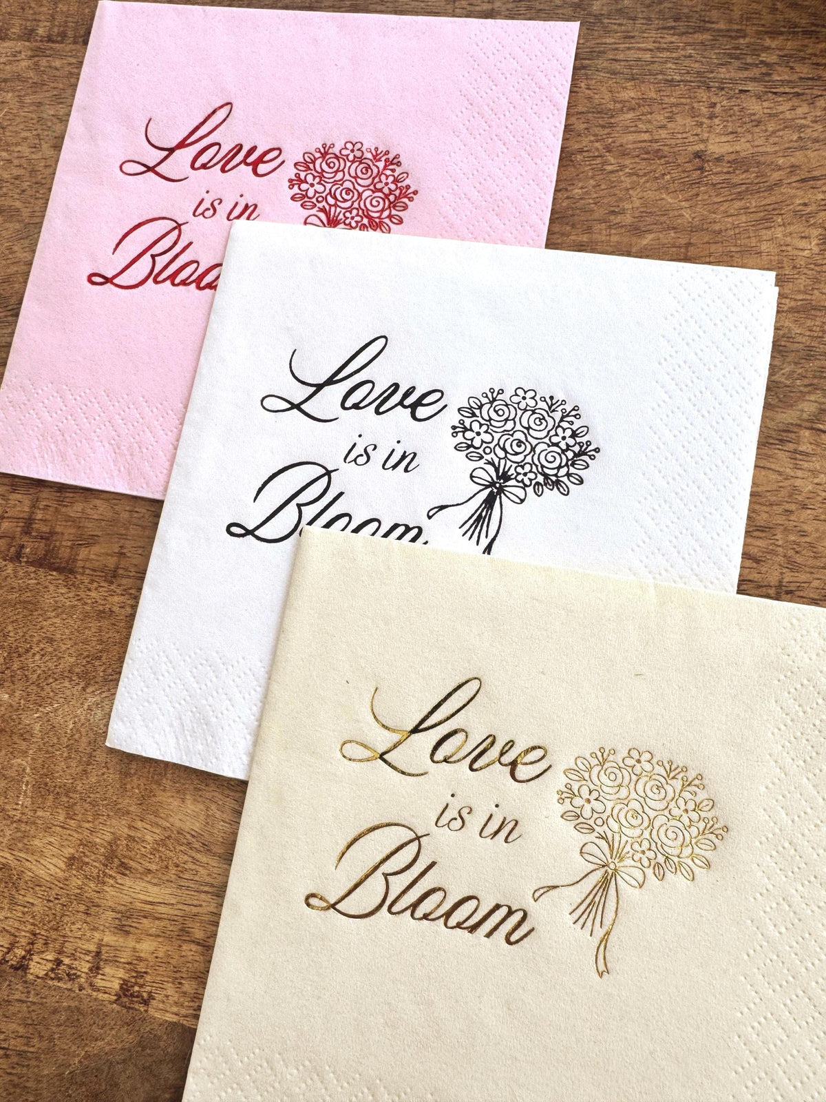 Three paper cocktail napkins in pink with red print, white with black print, and cream with gold print, each featuring the phrase Love is in Bloom and a floral bouquet.