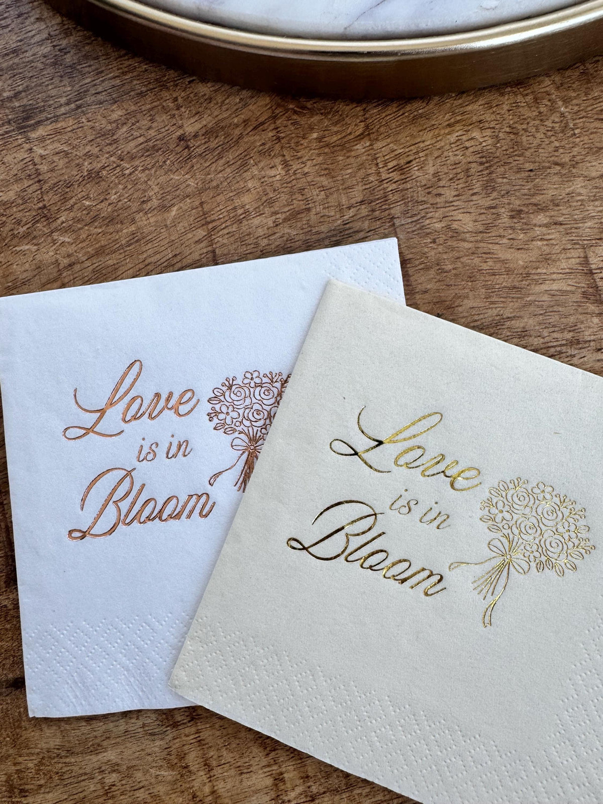 A white napkin with copper print and a cream napkin with gold print on a wood tabletop, displaying Love is in Bloom text and bouquet graphics.