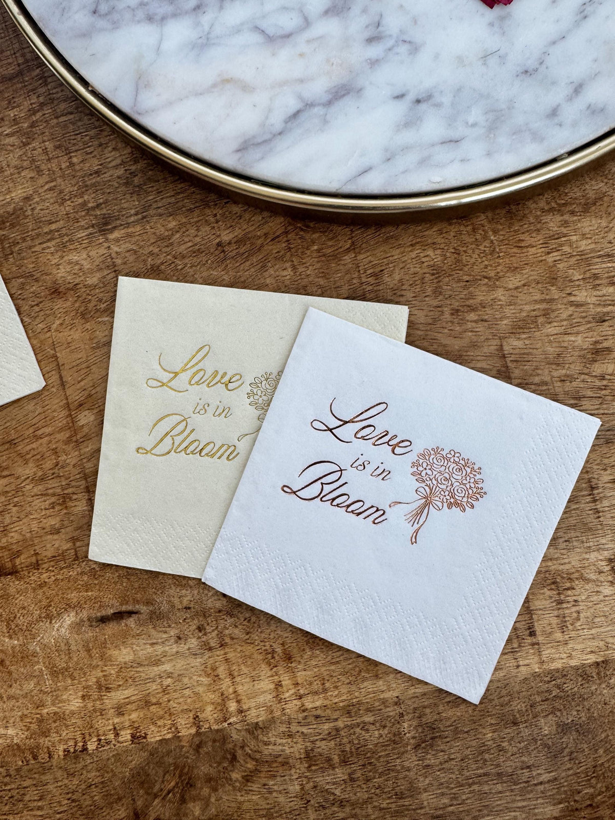 A cream napkin with gold foil and a white napkin with copper foil resting on a wooden surface next to a round tray.