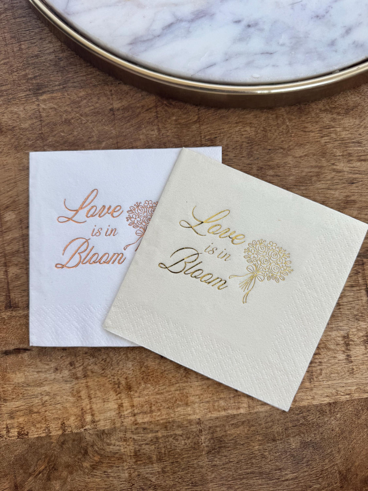 A white napkin with copper foil text and a cream napkin with gold foil text on a wooden background near a decorative tray.