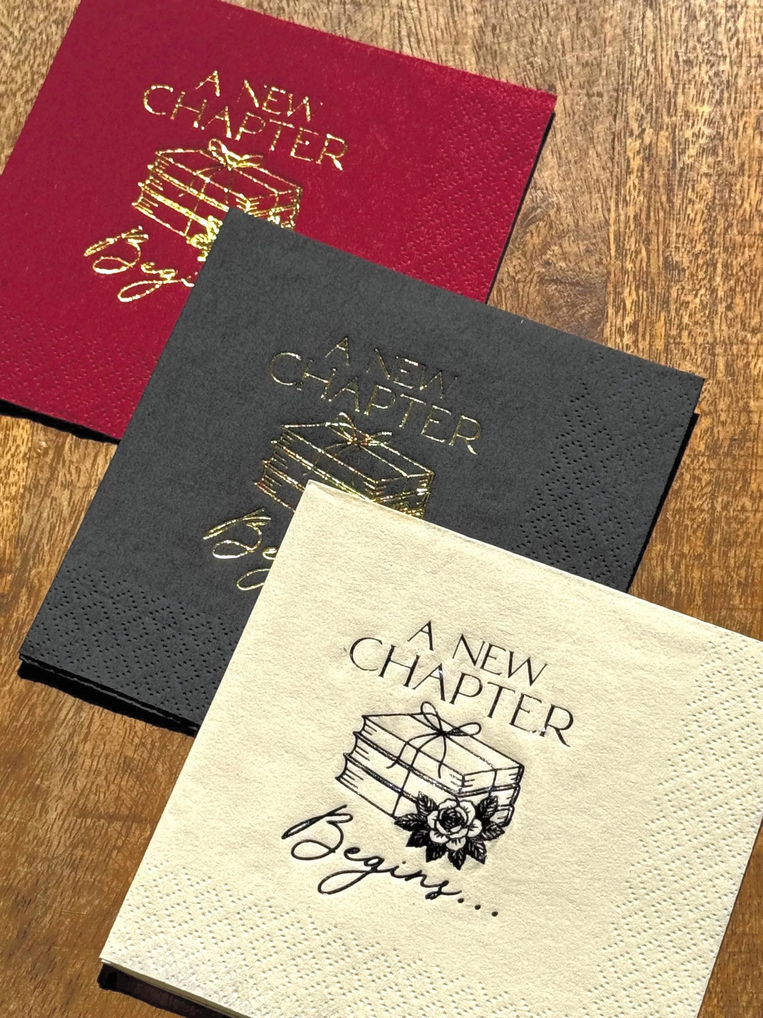 Foil-stamped “A New Chapter Begins” cocktail napkins in burgundy, black, and ivory, featuring a stacked book and floral design — perfect for baby showers, retirements, graduations, and book lover celebrations.