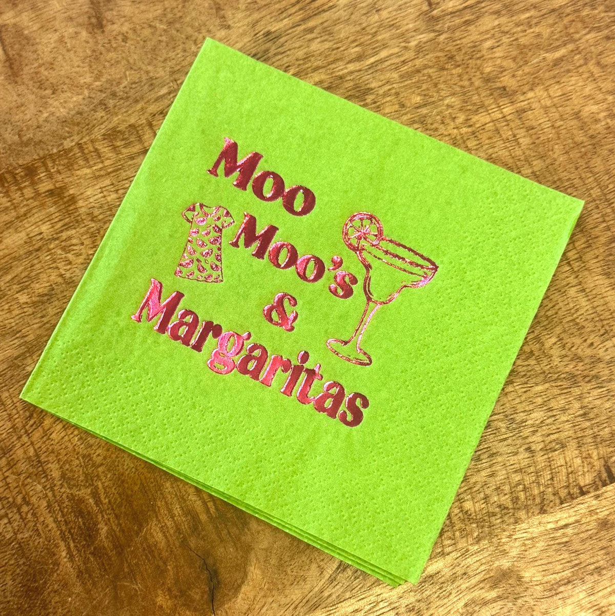 A stack of bright green cocktail napkins with the words "Moo Moo's & Margaritas" printed in pink. The design includes a margarita glass and a cow-print dress illustration.