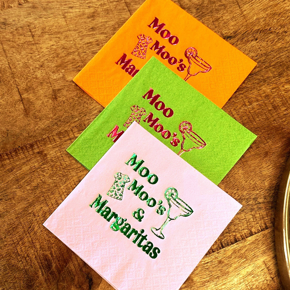 Three colorful cocktail napkins with the text "Moo Moo's & Margaritas". The napkins are orange, green, and pink, with a margarita glass and cow print design.