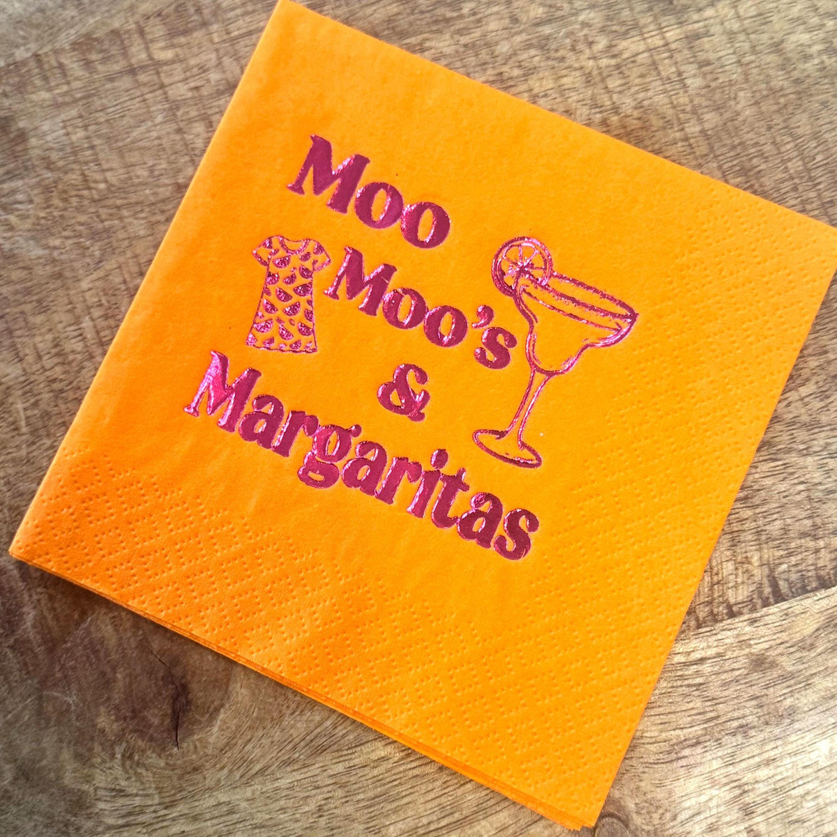 Orange cocktail napkin with the text "Moo Moo's & Margaritas" in pink. The napkin also features a pink illustration of a dress and a margarita glass.