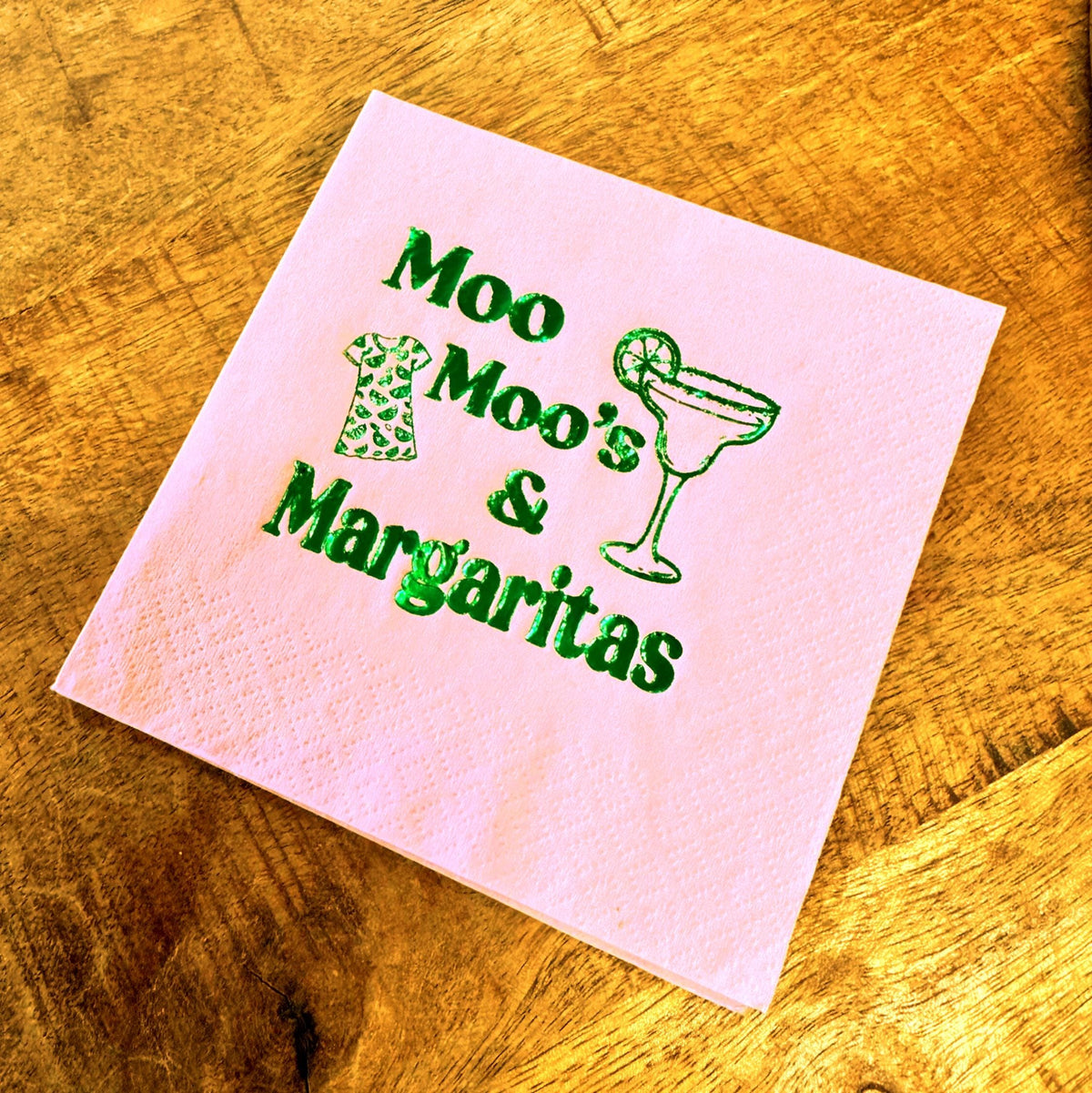 A pink paper napkin with the words "Moo Moo's & Margaritas" in green script. The napkin also features a green graphic of a dress and a margarita glass. The napkin is on a wooden surface.