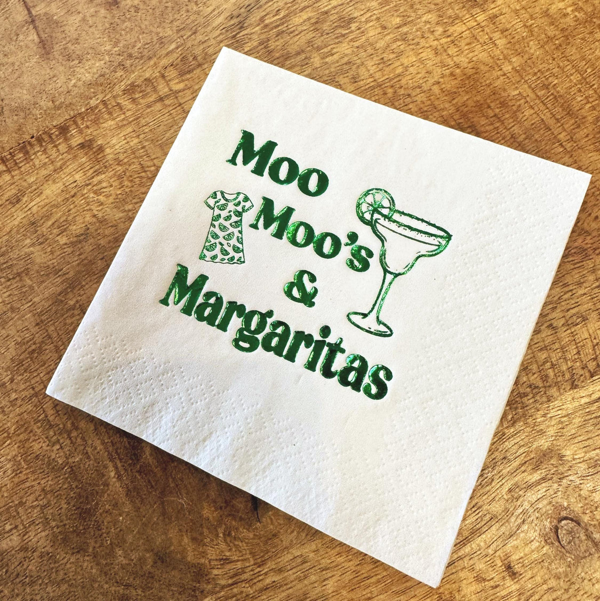 White paper cocktail napkin with the words "Moo Moo's & Margaritas" in green. The napkin also features a green illustration of a dress and a margarita glass with a lime wedge. The napkin is on a wooden surface.