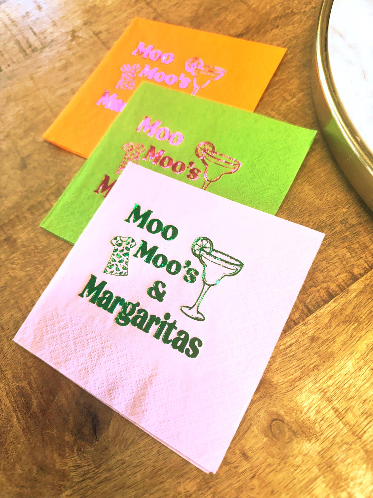 Three colorful cocktail napkins: orange, green, and pink. Each napkin features the text "Moo Moo's" and a margarita glass graphic. The pink napkin also includes the text "& Margaritas".