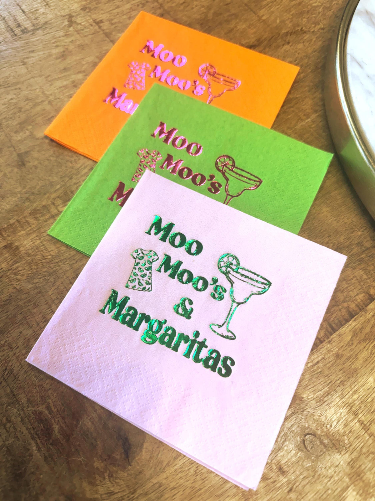 Three colorful cocktail napkins with the text "Moo Moo's & Margaritas". The napkins are orange, green, and pink, with a margarita glass and cow print design. The napkins are on a wooden surface.