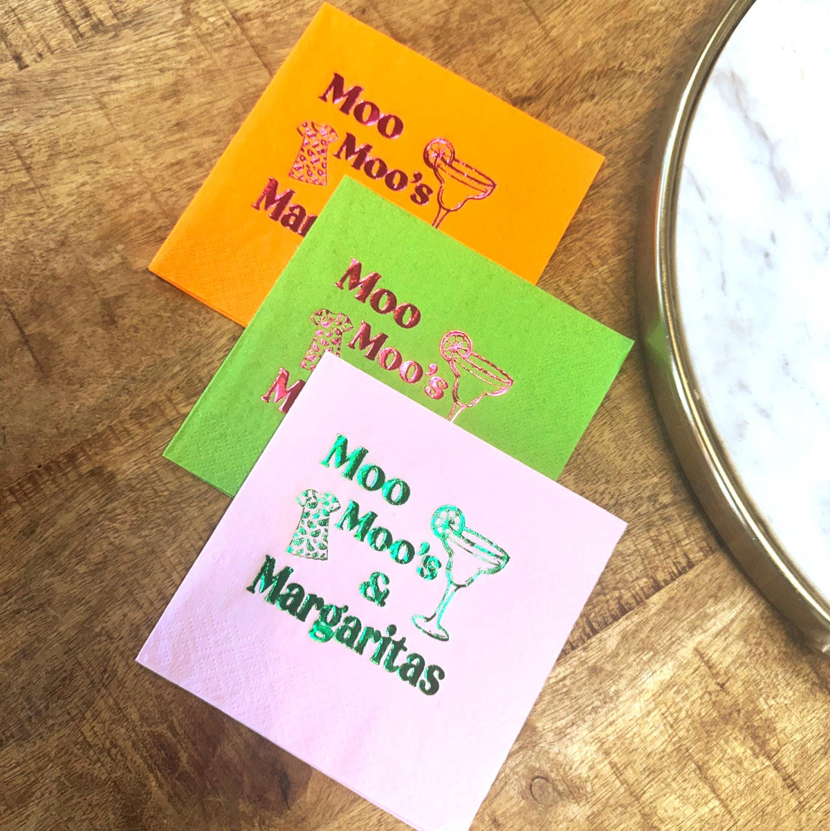 Three colorful cocktail napkins with the text "Moo Moo's & Margaritas". The napkins are orange, green, and pink, with a margarita glass and cow print design. The napkins are on a wooden surface.