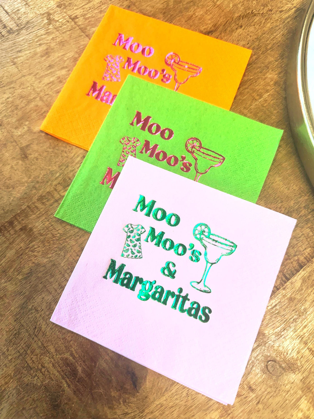 Three colorful cocktail napkins: orange, green, and pink. Each napkin features the text "Moo Moo's & Margaritas" with a margarita glass and a dress graphic. The napkins are arranged on a wooden surface.