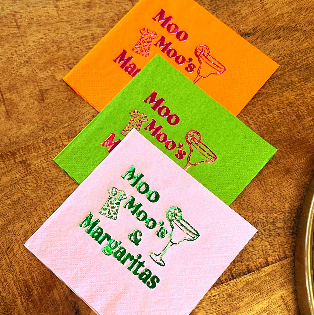 Three colorful cocktail napkins in orange, green, and pink, each with a different design. The napkins feature the text "Moo Moo's & Margaritas" and margarita glass illustrations. The napkins are on a wooden surface.